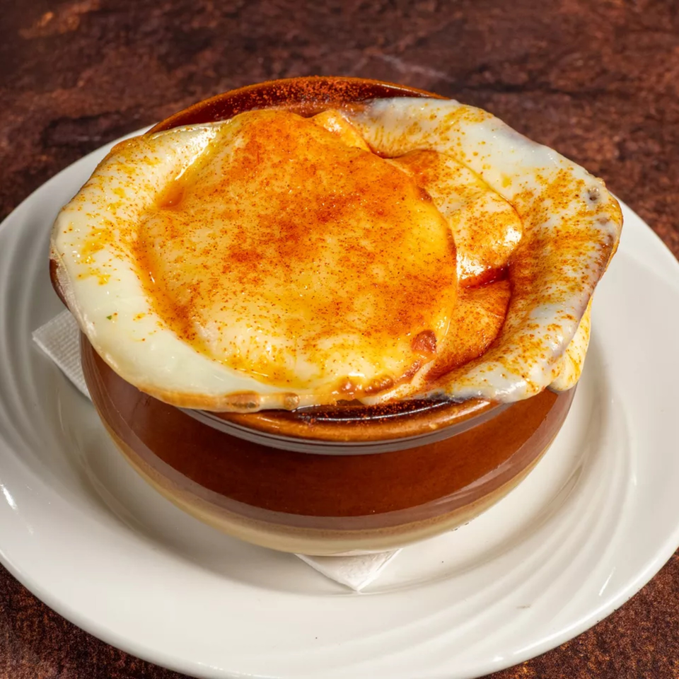 A brown ceramic bowl filled with a creamy dish topped with melted cheese and paprika, on a white plate.